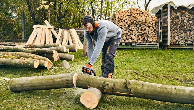 A man wearing hearing protection cuts a large log with a STIHL chainsaw in a backyard filled with chopped firewood.