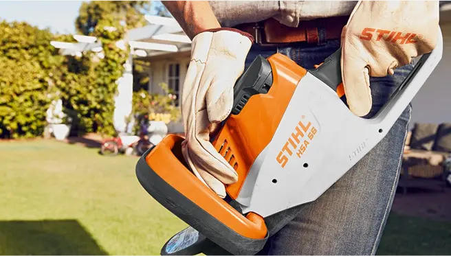 A close-up of a person holding a STIHL HSA 56 hedge trimmer, with focus on the tool and gloves, in a residential garden setting.