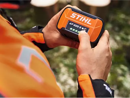 A person holds a STIHL AP 500 S battery, showing its charge level with LED indicators, while wearing an orange work jacket outdoors.