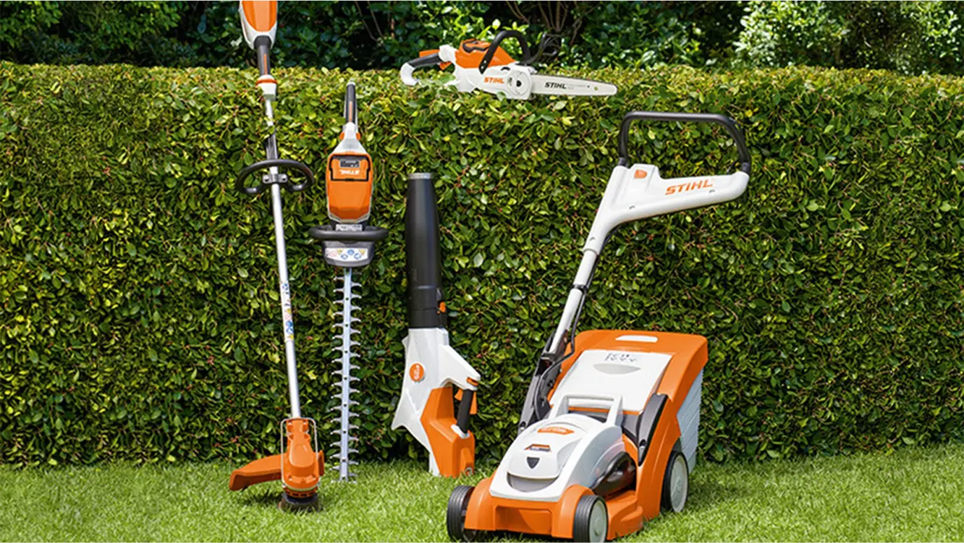 A selection of STIHL battery-powered garden tools displayed on a neatly trimmed lawn in front of a tall green hedge, including a lawn mower, hedge trimmer, leaf blower, and chainsaw.