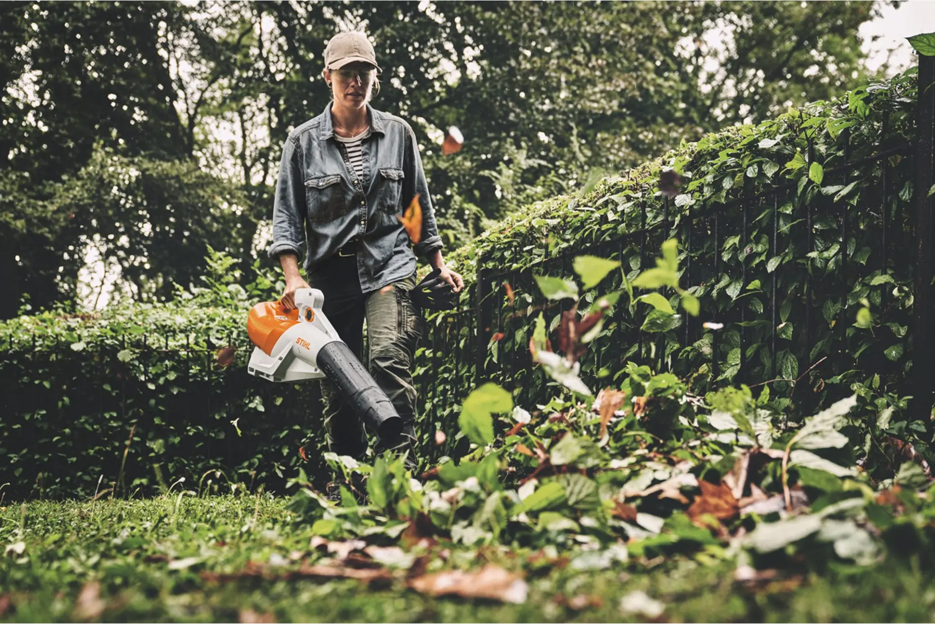 A woman in workwear uses a STIHL leaf blower to clear fallen leaves and garden debris in a lush, green backyard with dense shrubbery and trees.