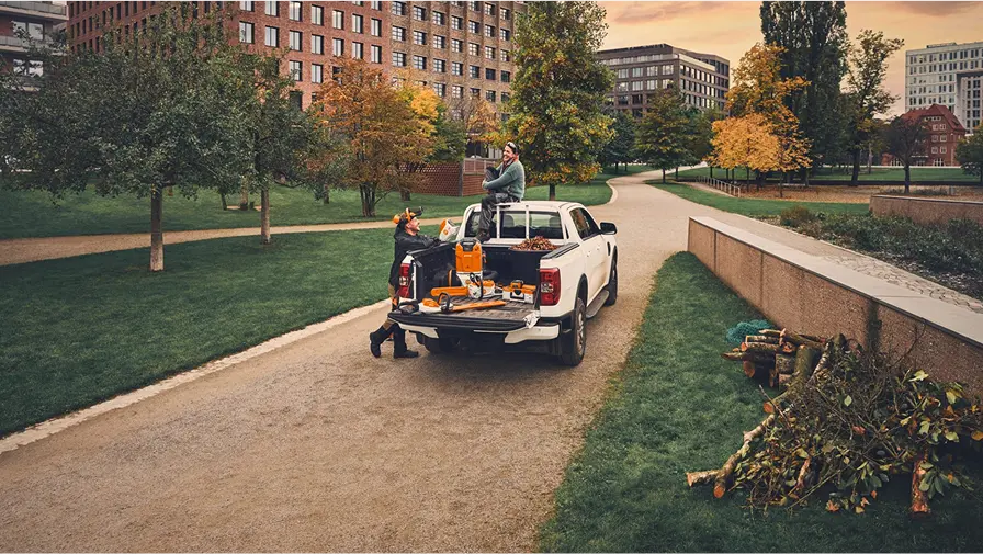 Two people load STIHL garden tools onto the back of a white pickup truck parked on a path in a landscaped urban park, with autumn trees and office buildings in the background.