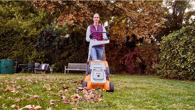A woman pushes a STIHL lawn mower across a leaf-covered lawn in a garden surrounded by trees during autumn.